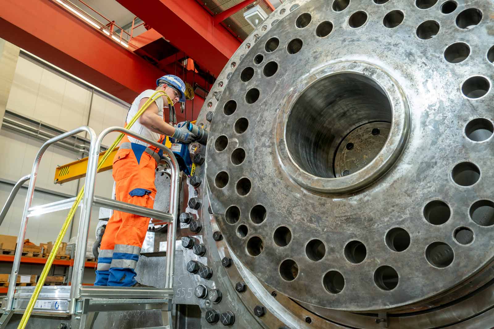 Eine Person arbeitet an einer großen Metallstruktur mit vielen Löchern. Sicherheitsausrüstung und Gerüst sind sichtbar.