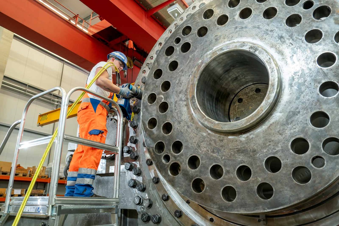 A person is working on a large metal structure with numerous holes. Safety equipment and scaffolding are visible.