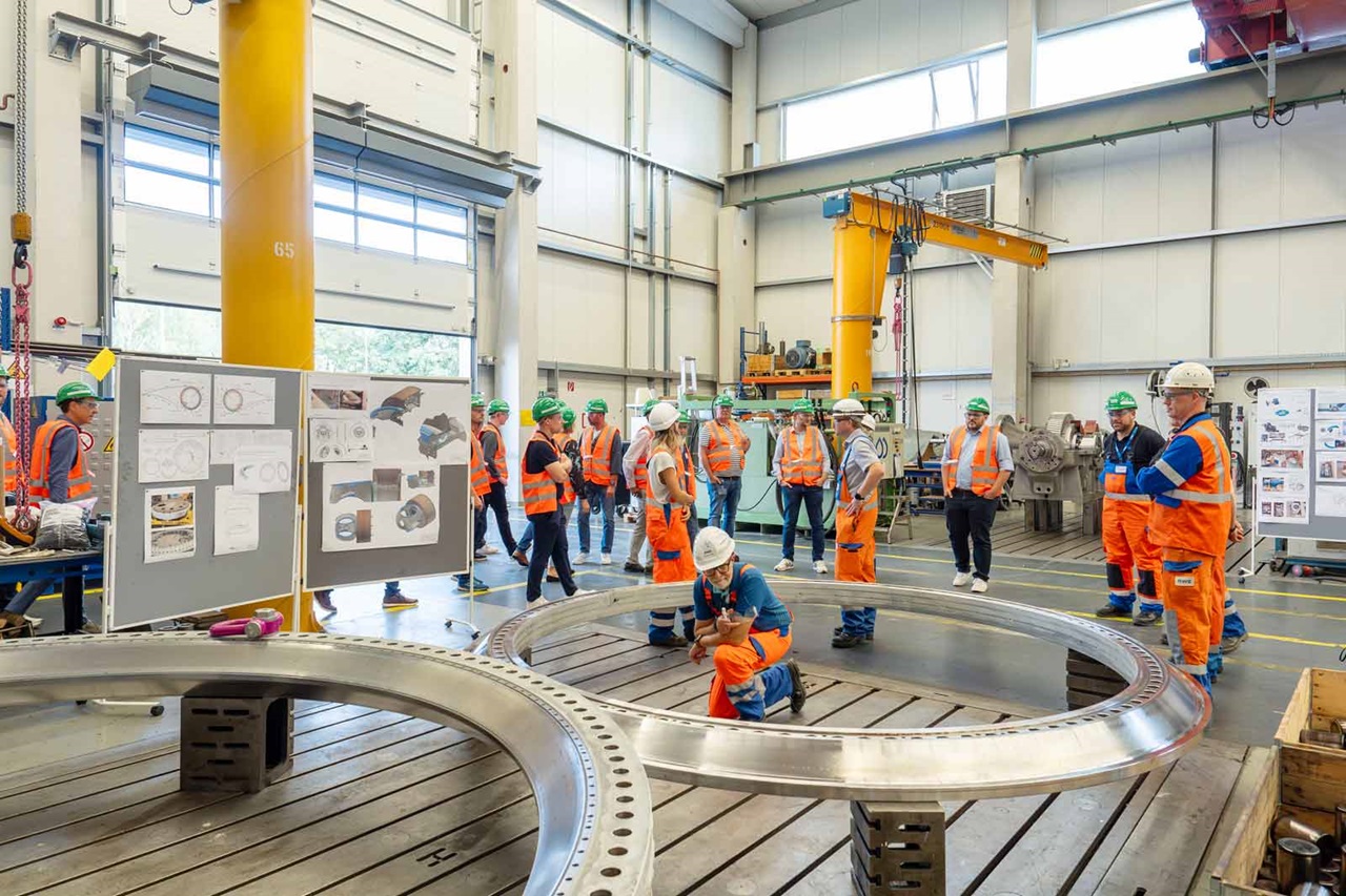 A group of professionals in orange safety vests and helmets discusses in front of a large metal component.