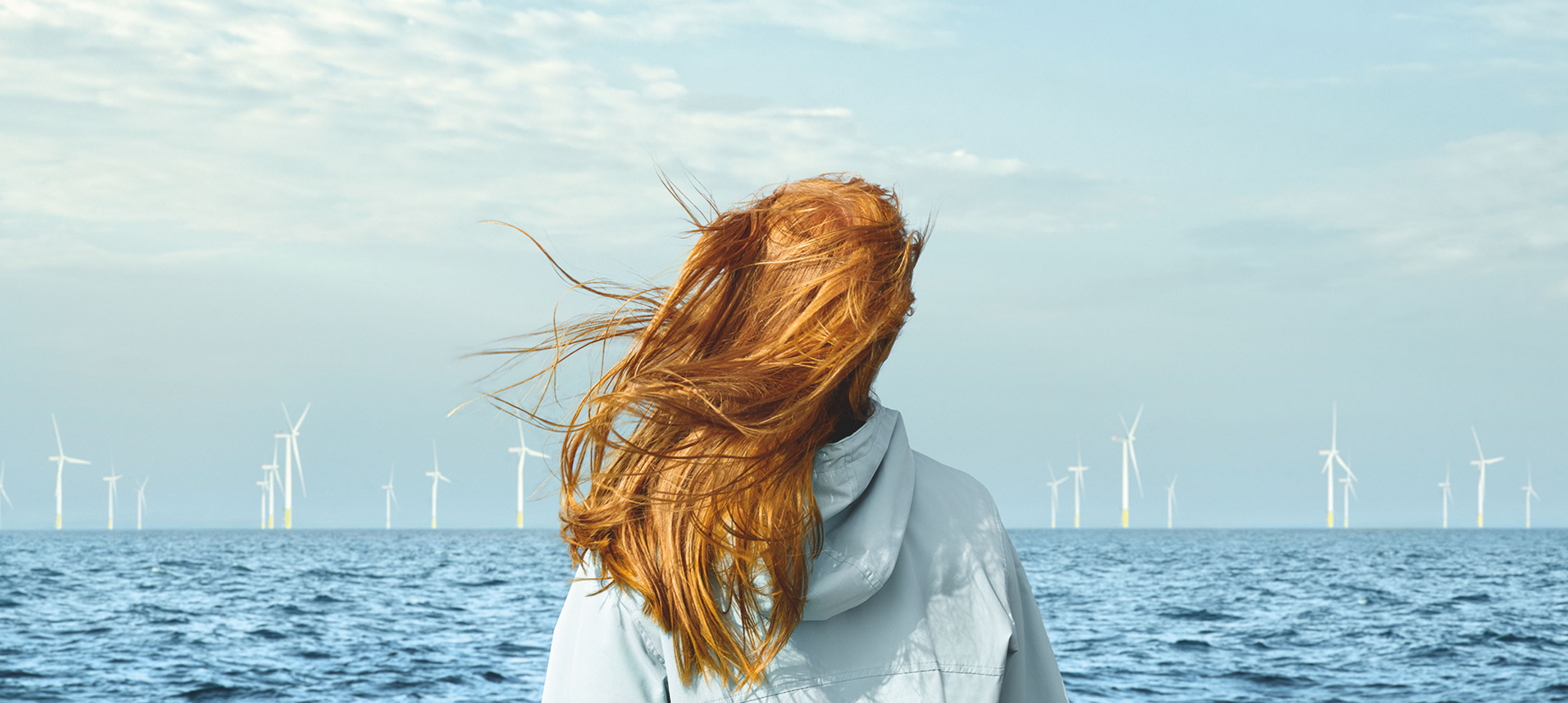 Eine Frau mit langen roten Haaren steht am Wasser und schaut auf Windkraftanlagen im Hintergrund unter einem bewölkten Himmel.