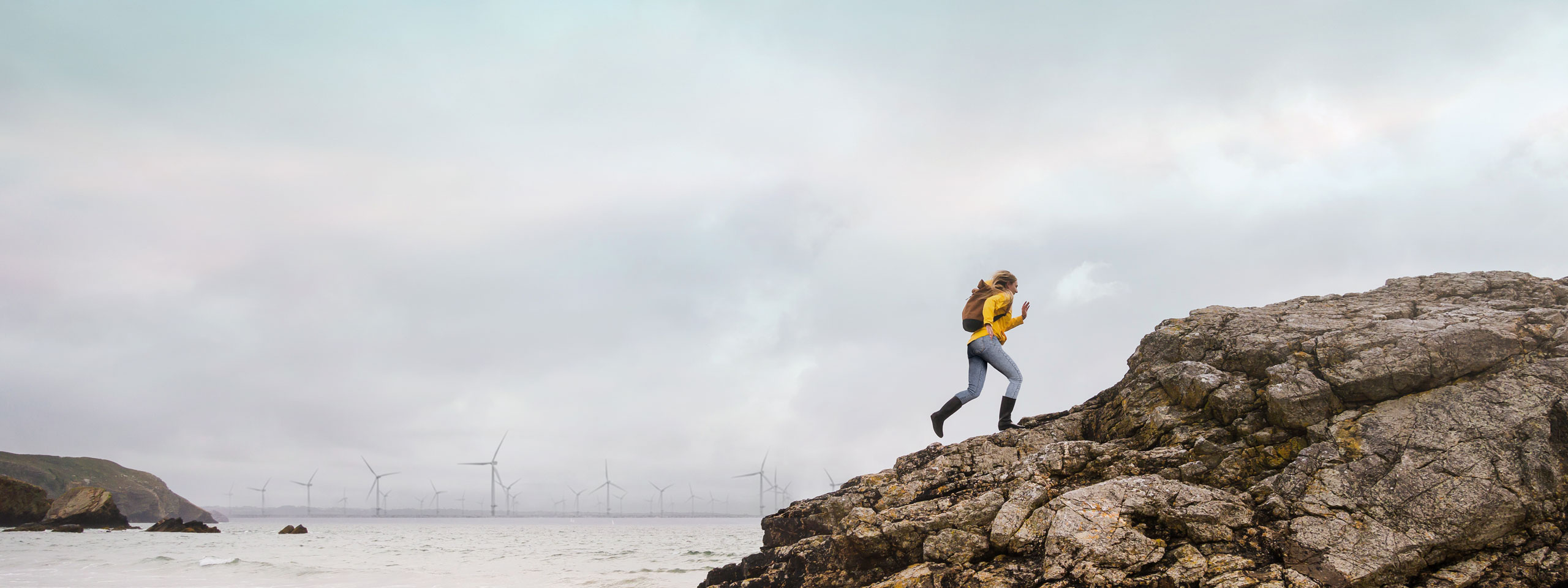 A woman in a yellow jacket climbs a rocky shore with wind turbines visible in the background under a cloudy sky.