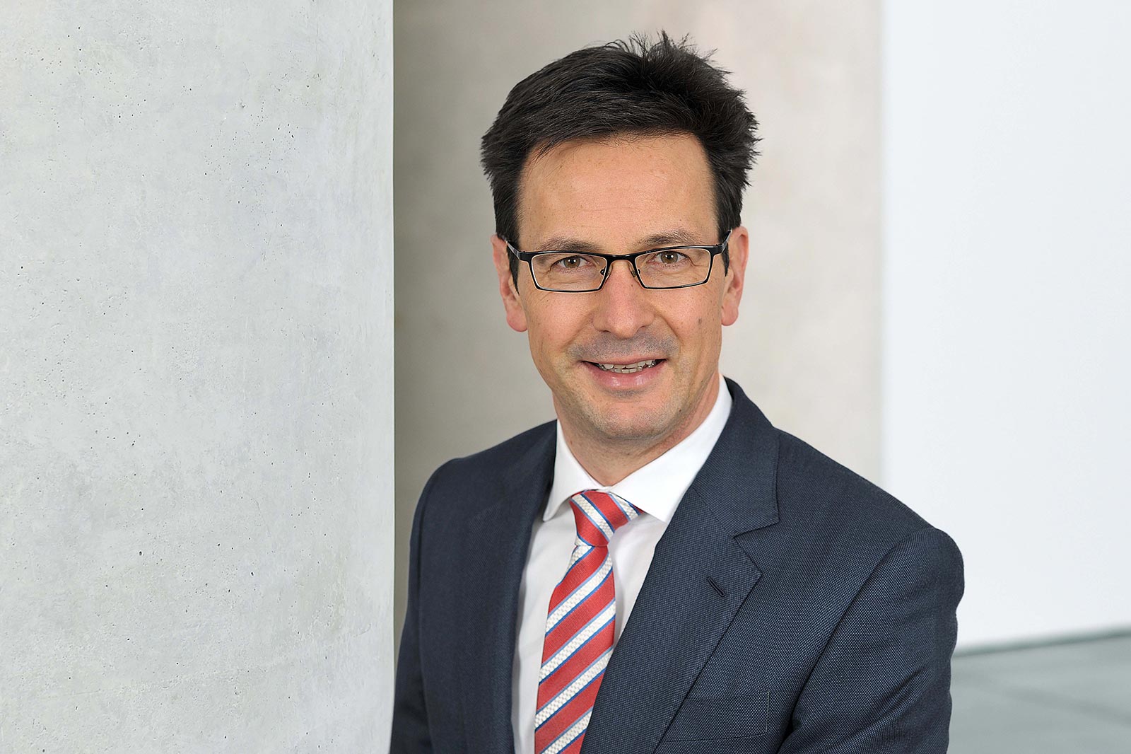 Ein Mann in einem Anzug mit gestreifter Krawatte steht neben einer hellen, strukturierten Wand. Portrait von Bernhard Günther.