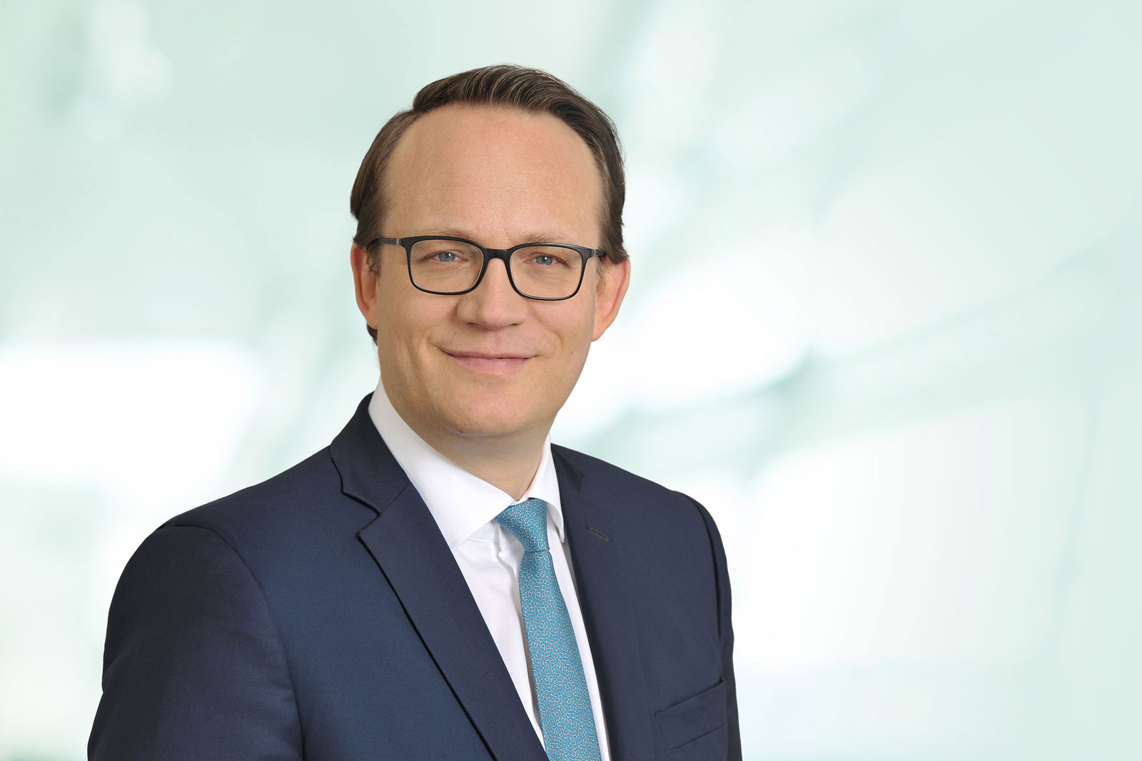 A man in a suit with a tie stands against a bright, blurred background. Portrait of Dr. Markus Krebber.