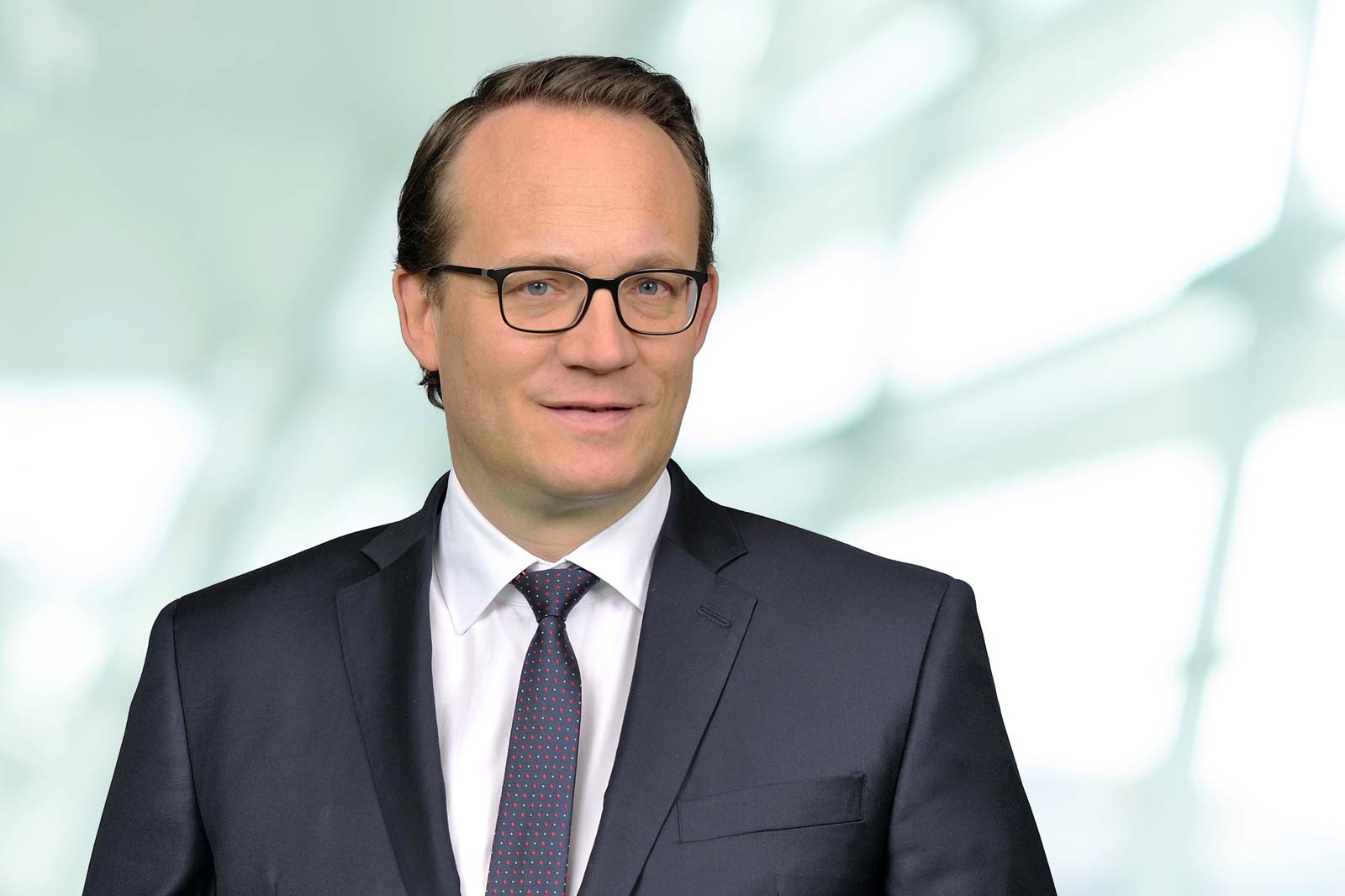 A man in a suit and tie stands before a blurred, bright background. Portrait of Dr. Markus Krebber.