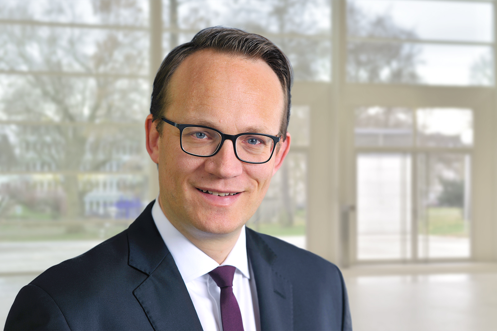 Ein Mann in einem Anzug steht vor einer modernen Fensterfront. Der Hintergrund ist hell und freundlich. Portrait von Dr. Markus Krebber.
