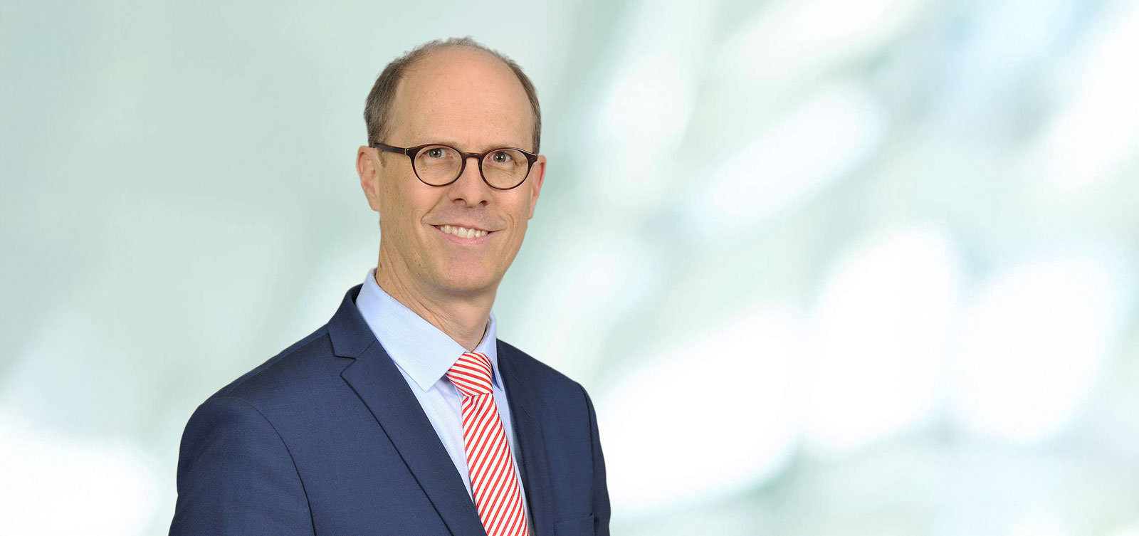 Ein Mann in einem blauen Anzug mit einem rot-weiß gestreiften Krawatte steht vor einem hellen, verschwommenen Hintergrund. Portrait von Dr. Michael Müller.