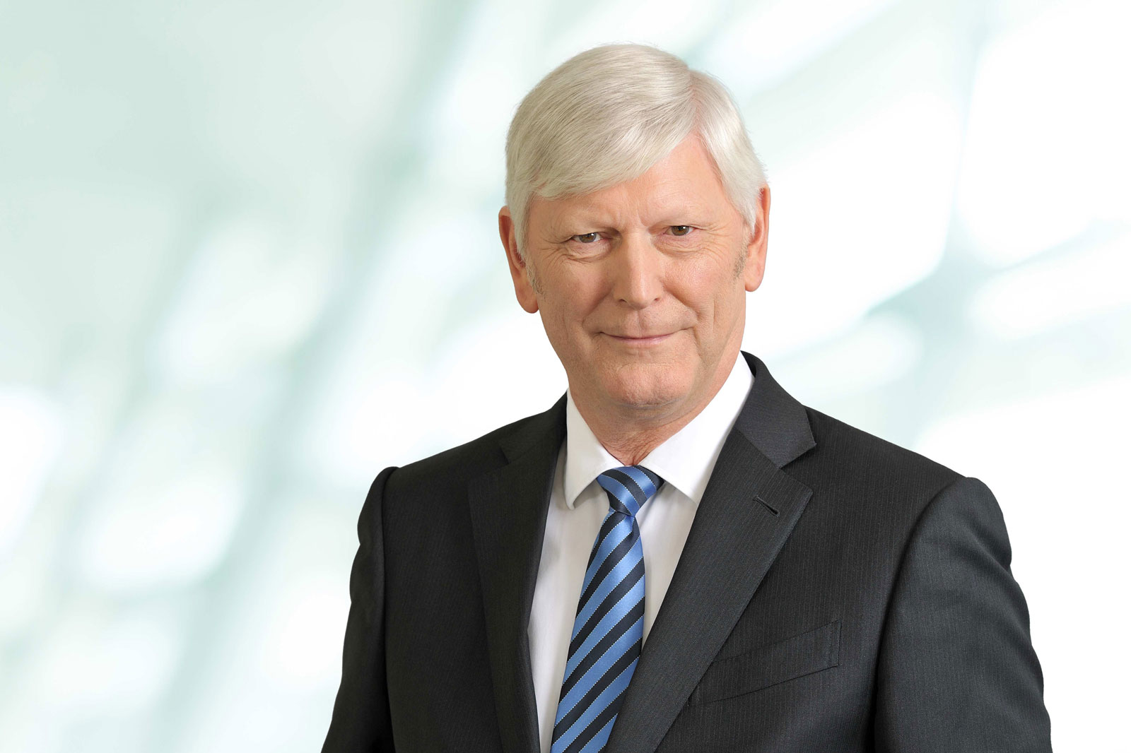 Ein älterer Mann in einem Anzug mit blauen Streifen auf der Krawatte vor einem hellen, verschwommenen Hintergrund. Portrait von Rolf Martin Schmitz.