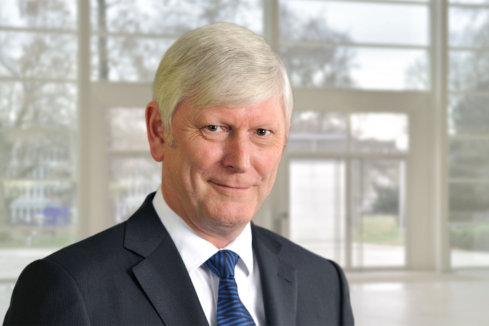 A man in a dark blue suit with a striped tie stands in a modern, bright room. Portrait of Rolf Martin Schmitz.