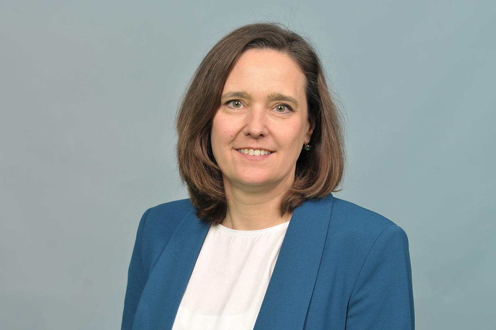 Eine Frau mit schulterlangen braunen Haaren trägt einen blauen Blazer und eine weiße Bluse vor einem hellgrauen Hintergrund. Portrait von Andrea Rodemann.