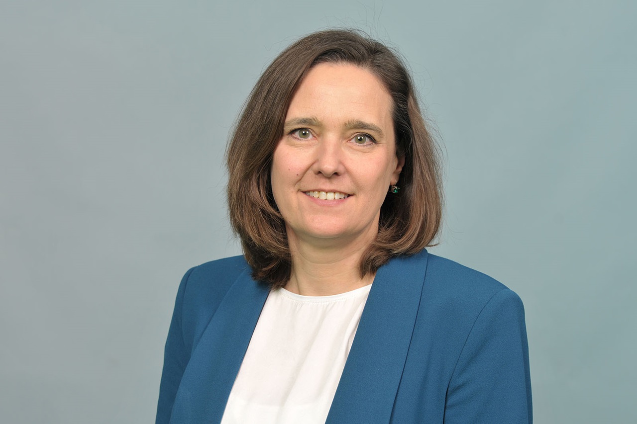 Eine Frau mit schulterlangen braunen Haaren trägt einen blauen Blazer und eine weiße Bluse vor einem hellgrauen Hintergrund. Portrait von Andrea Rodemann.