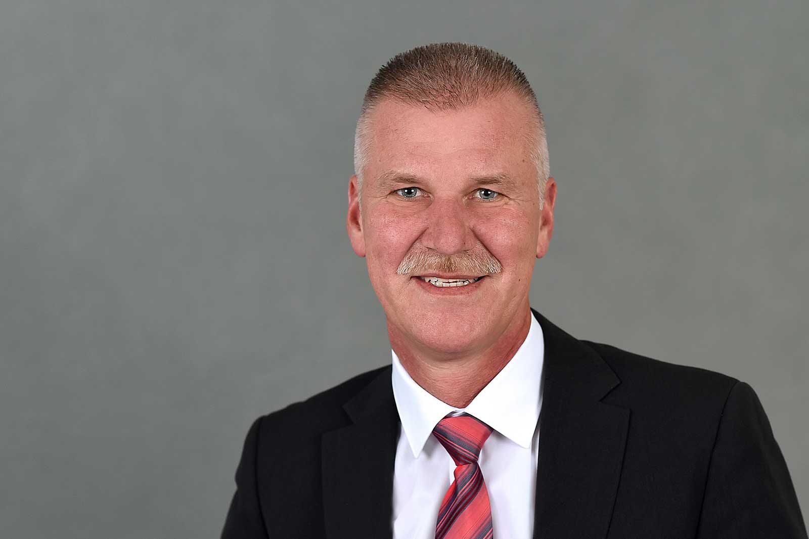 A man in a black suit with a striped tie stands in front of a grey background. Portrait of Michael Bochinsky.