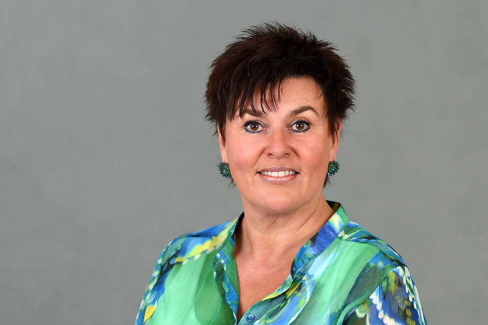 A woman with short dark hair wears a colourful shirt and green earrings against a grey background. Portrait of Sandra Bossemeyer.