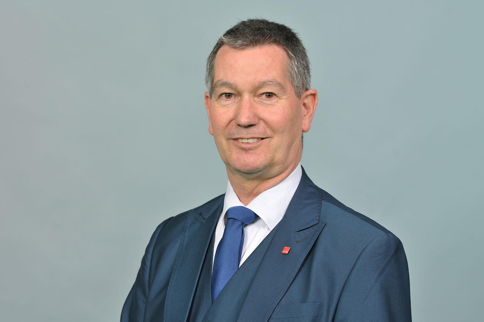 A person with short gray hair wearing a dark navy suit, white shirt, and blue tie stands against a pale blue-gray studio background. Portrait of Christoph Schmitz-Dethlefsen.
