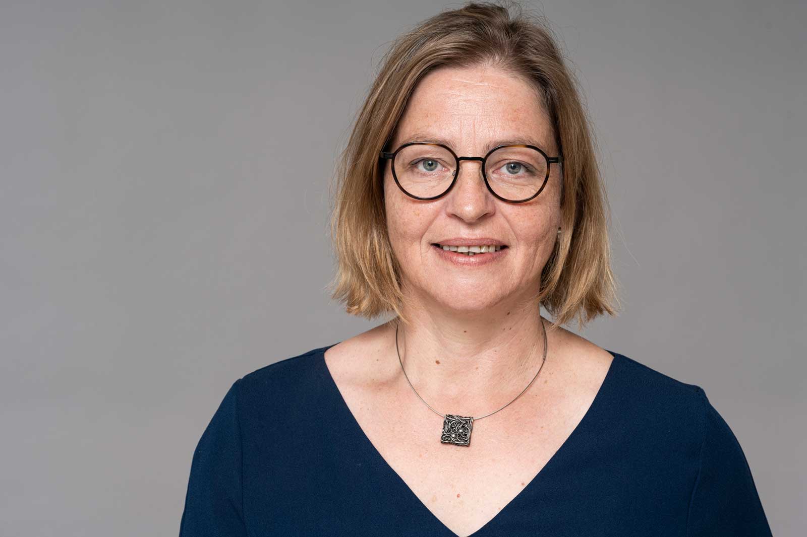 The person wears a dark top with a V-neck and a silver necklace featuring a square pendant. Portrait of Dagmar Paasch.