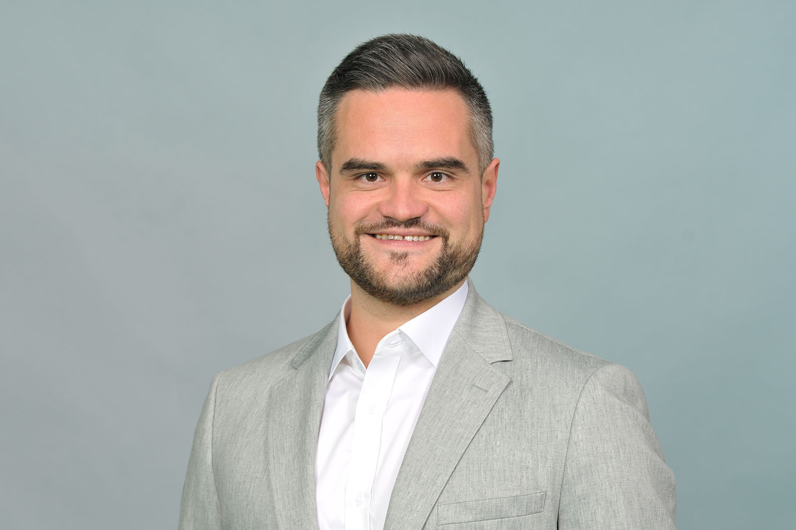 Man with short dark hair wearing a light grey suit jacket and white shirt against a pale grey background. Portrait of Daniel Kober.