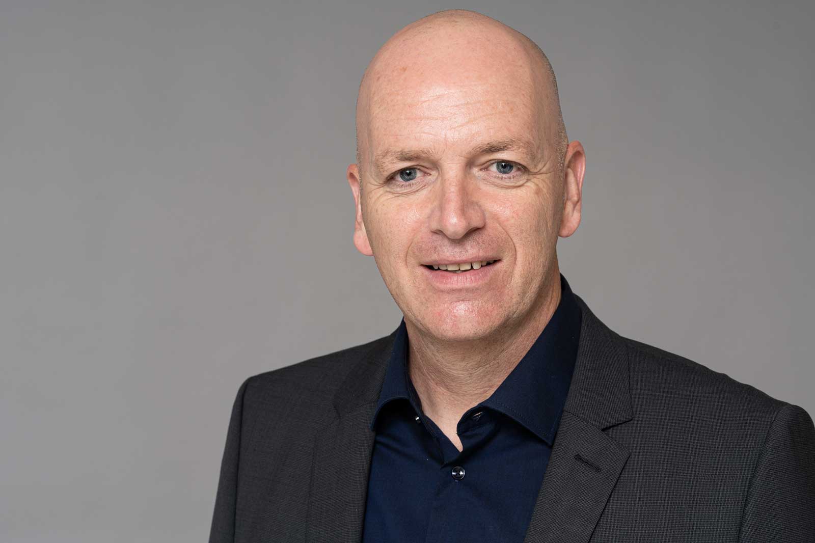 A man in a dark suit wearing a black shirt against a grey background. Portrait of Dirk Schumacher.