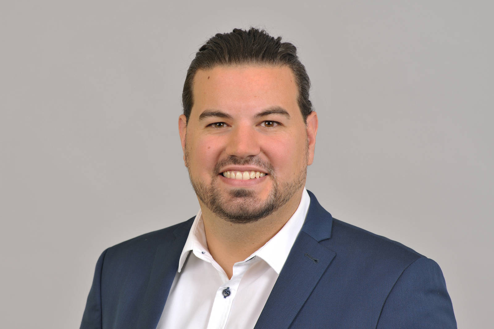 A man with dark, tied-back hair is wearing a blue suit and a white shirt. The background is grey. Portrait of Matthias Dürbaum.