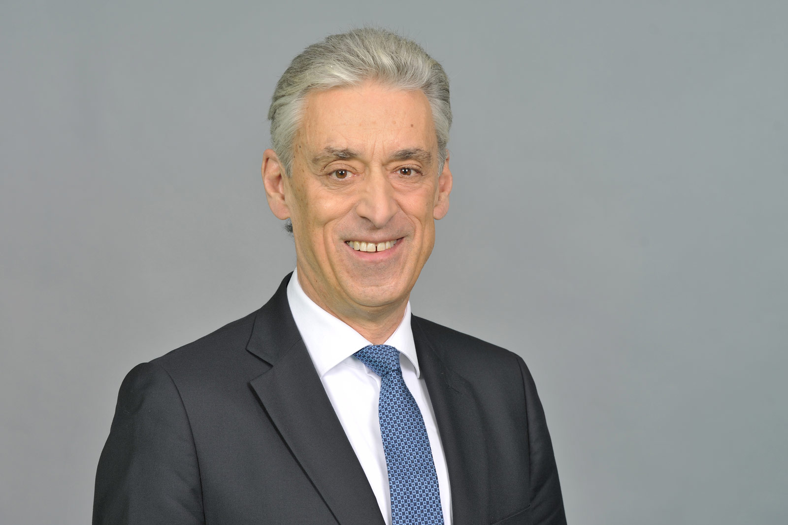 A man in a black suit with a blue tie against a grey background. Portrait of Dr Frank Appel.