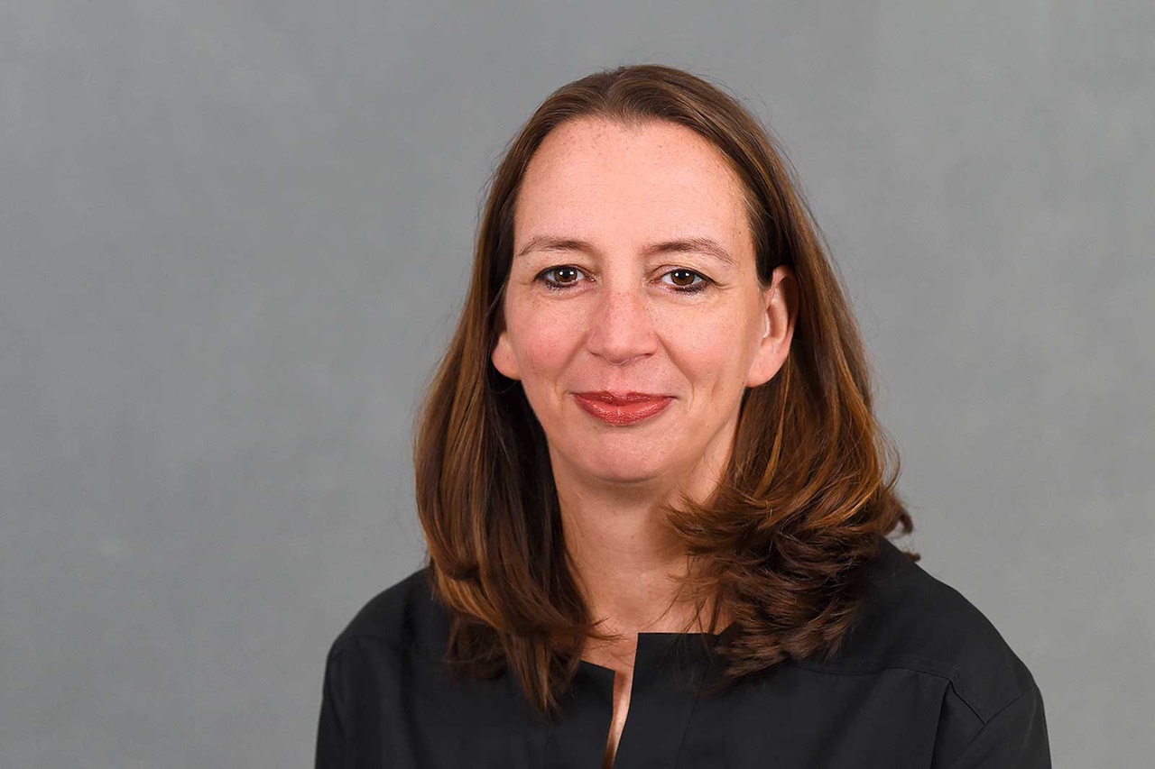 Eine Person mit langen, brünetten Haaren trägt ein schwarzes Oberteil. Der Hintergrund ist grau. Portrait von Ute Gerbaulet.