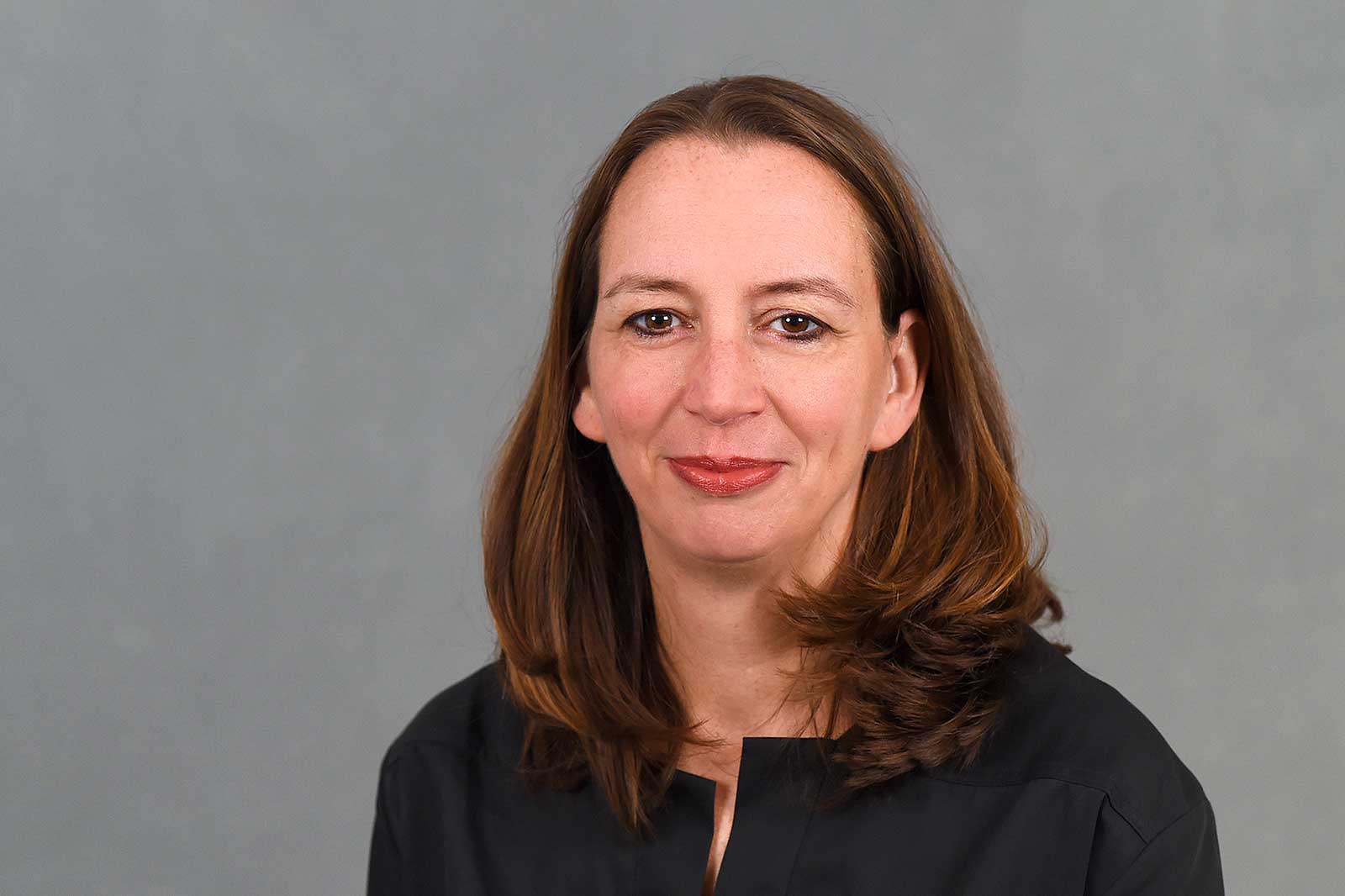 A person with long, brown hair is wearing a black top. The background is grey. Portrait of Ute Gerbaulet.