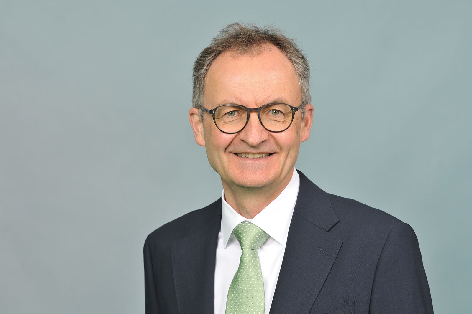 Man in a dark navy suit, white shirt and pale green tie stands against a blue-gray background. Portrait of Gordon Dreyer.