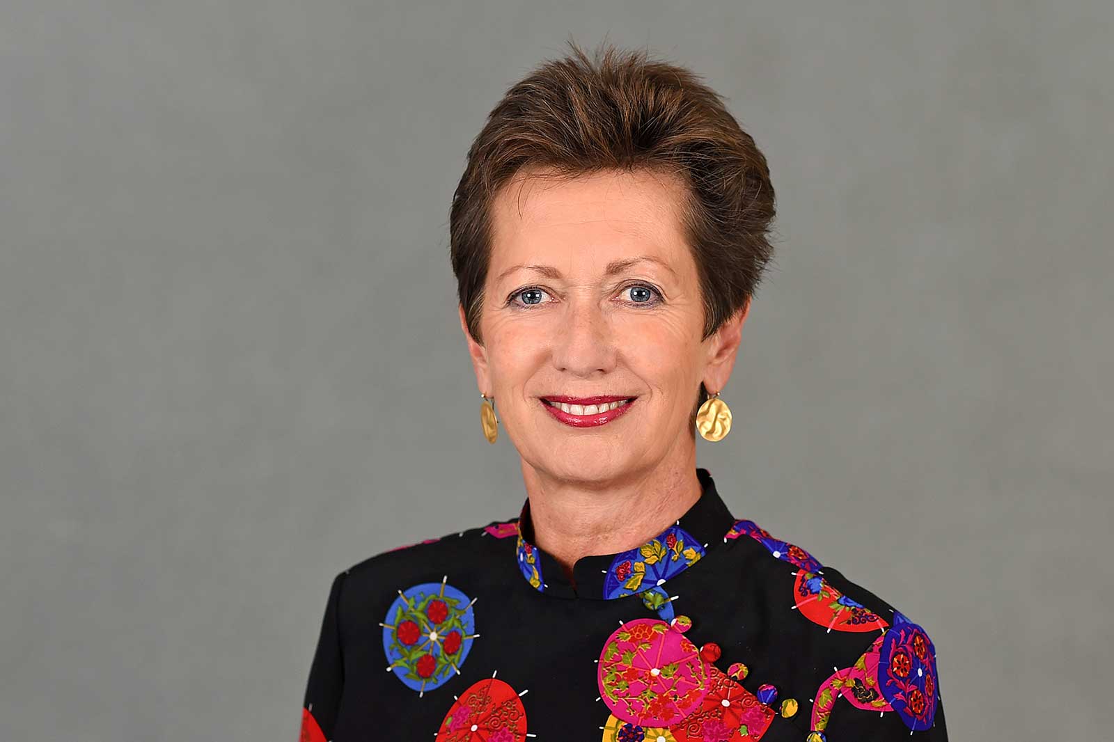 Eine Person trägt ein buntes Kleid mit Blumenmustern vor einem grauen Hintergrund. Portrait von Mag. Dr. h.c. Monika Kircher.