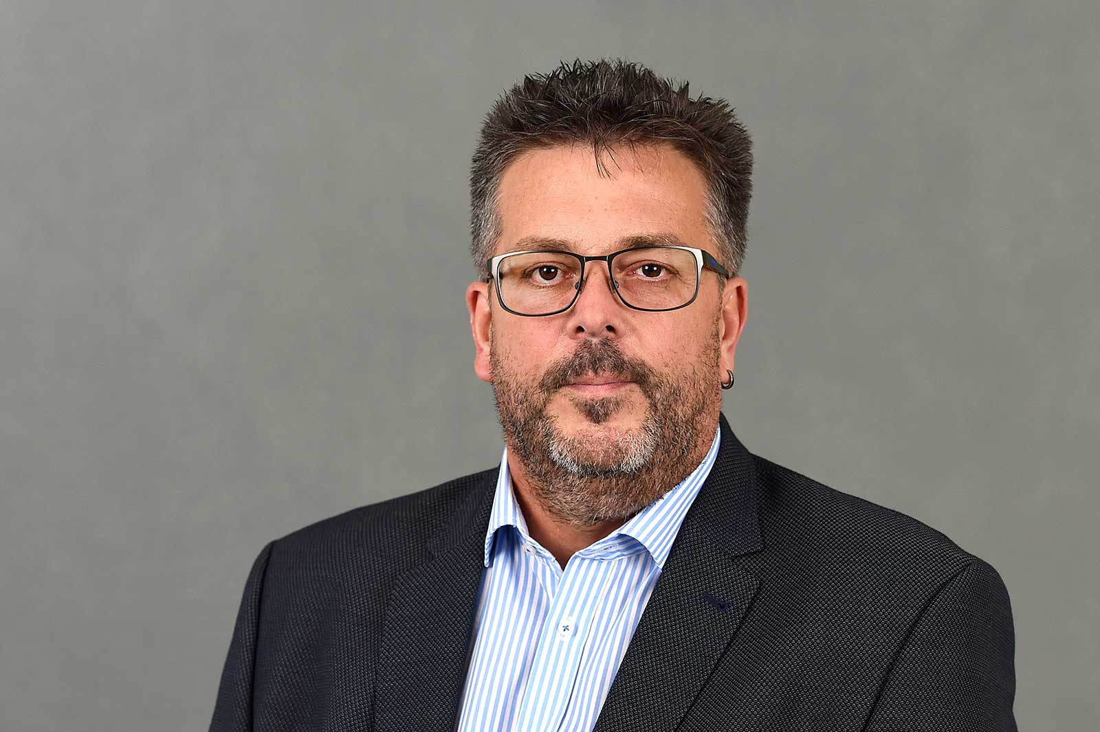 Ein Mann in einem Anzug mit einem karierten Hemd steht vor einem grauen Hintergrund. Portrait von Harald Louis.