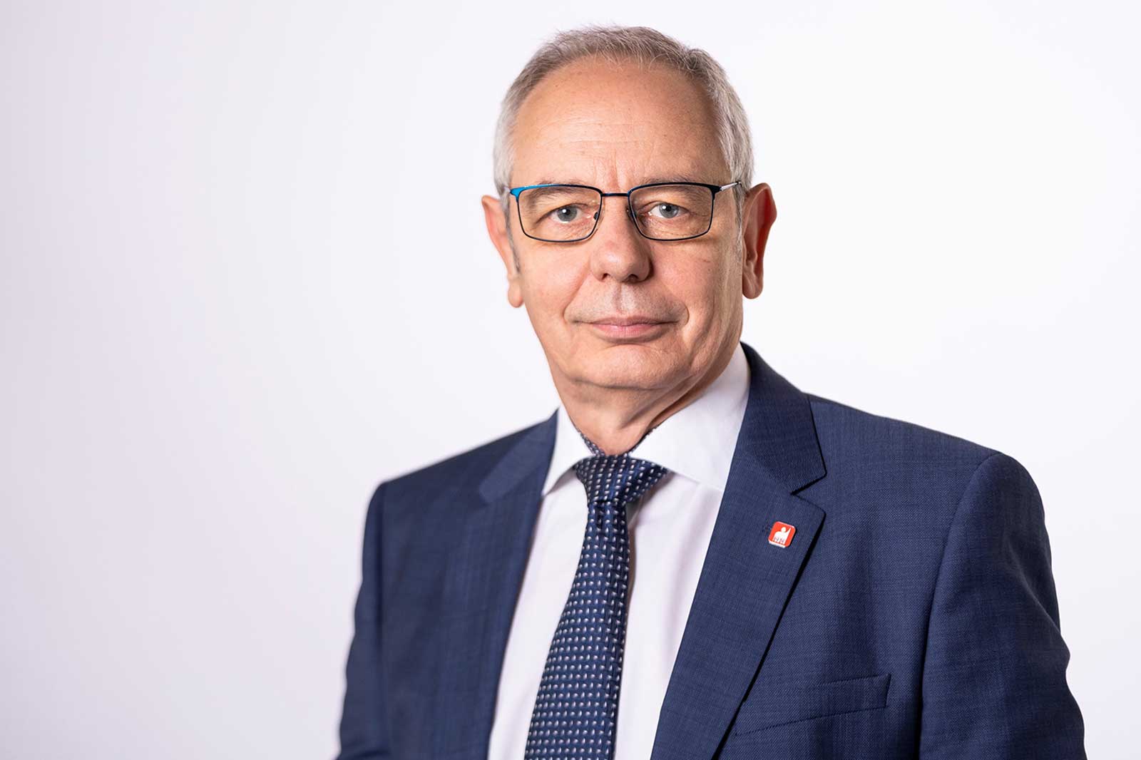 Ein Mann in einem dunkelblauen Anzug mit einer gestreiften Krawatte und einem roten Emblem auf dem Revers vor einem weißen Hintergrund. Portrait von Michael Vassiliadis.