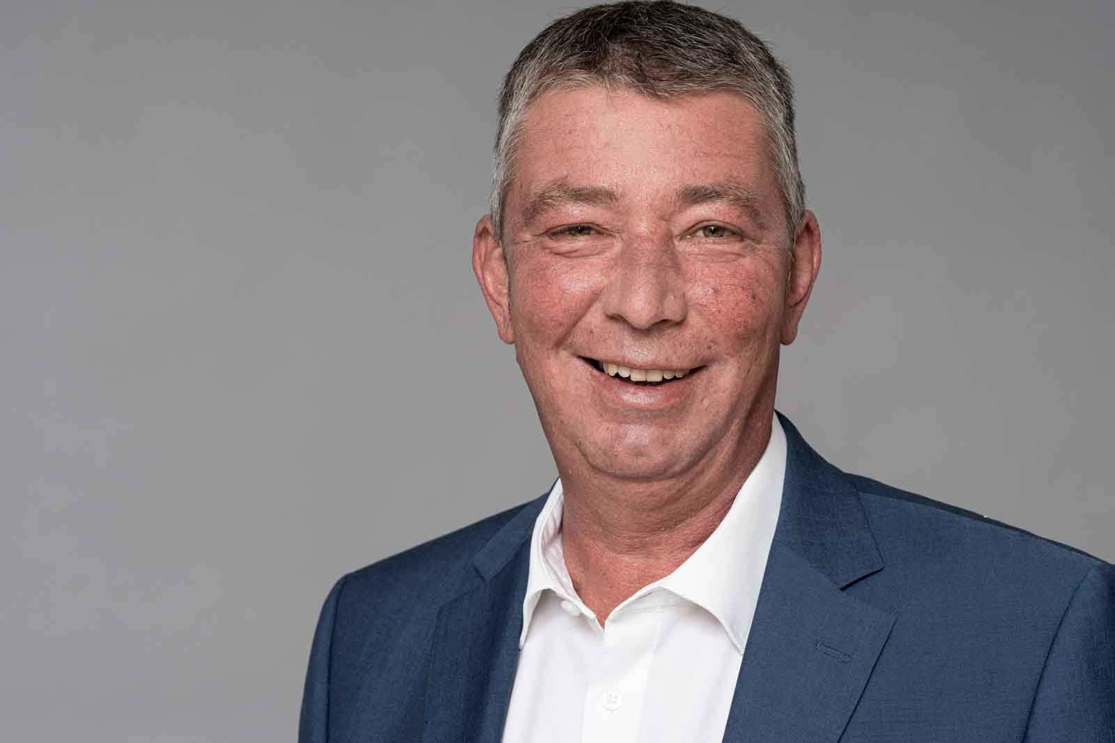 A man in a suit with a shirt stands against a grey background. He has grey hair and is wearing a blue suit. Portrait of Reiner van Limbeck.