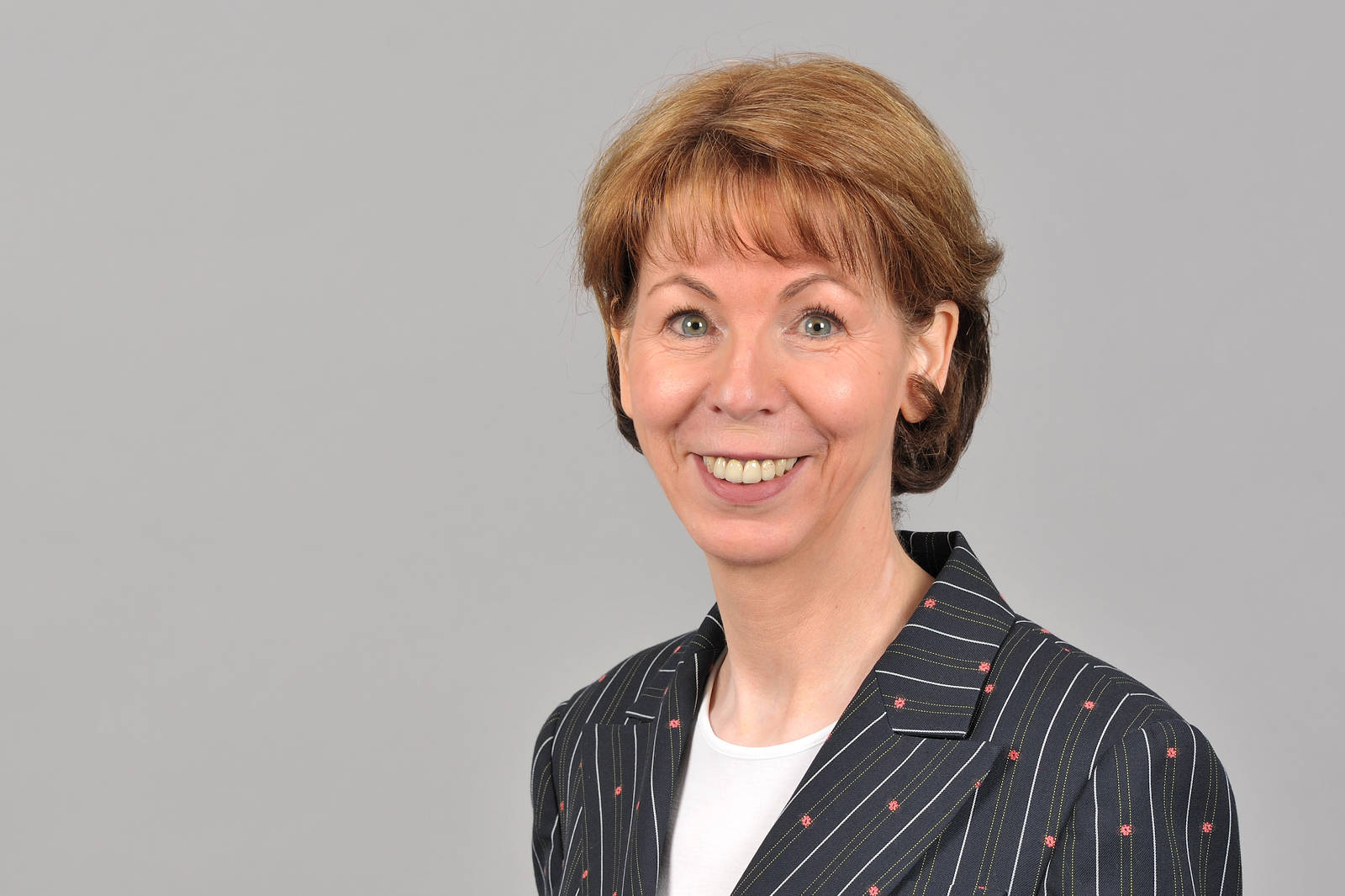 A person with brown hair is wearing a striped blazer and stands against a grey background. Portrait of Hauke Stars.