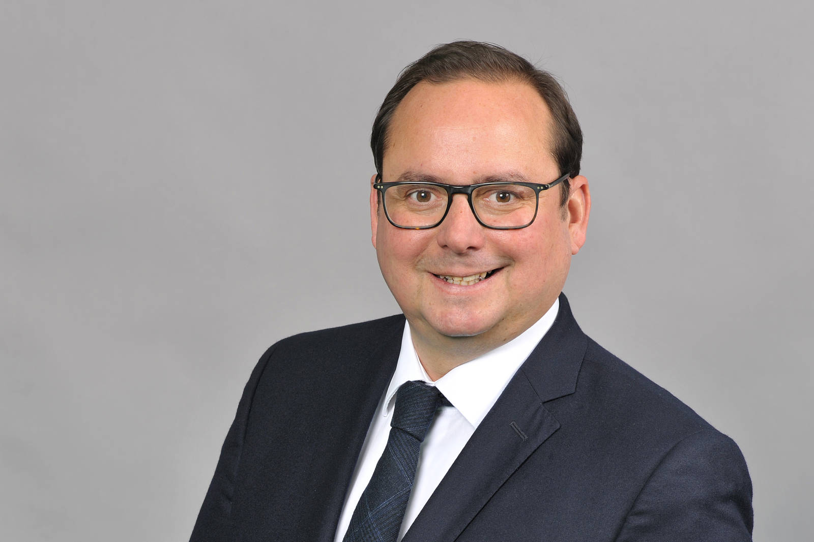 Ein Mann in einem dunklen Anzug und einer Krawatte, vor einem grauen Hintergrund. Er hat braunes Haar und trägt einen weißen Hemdkragen. Portrait von Thomas Kufen.