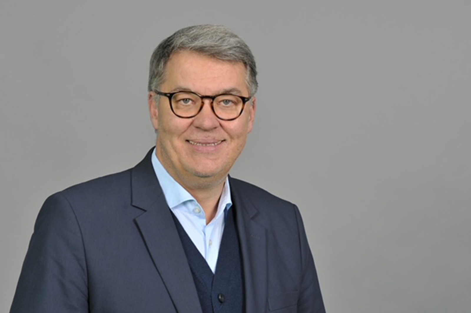 Ein Mann in einem dunklen Anzug und einer hellen Bluse steht vor einem grauen Hintergrund. Portrait von Thomas Westphal.