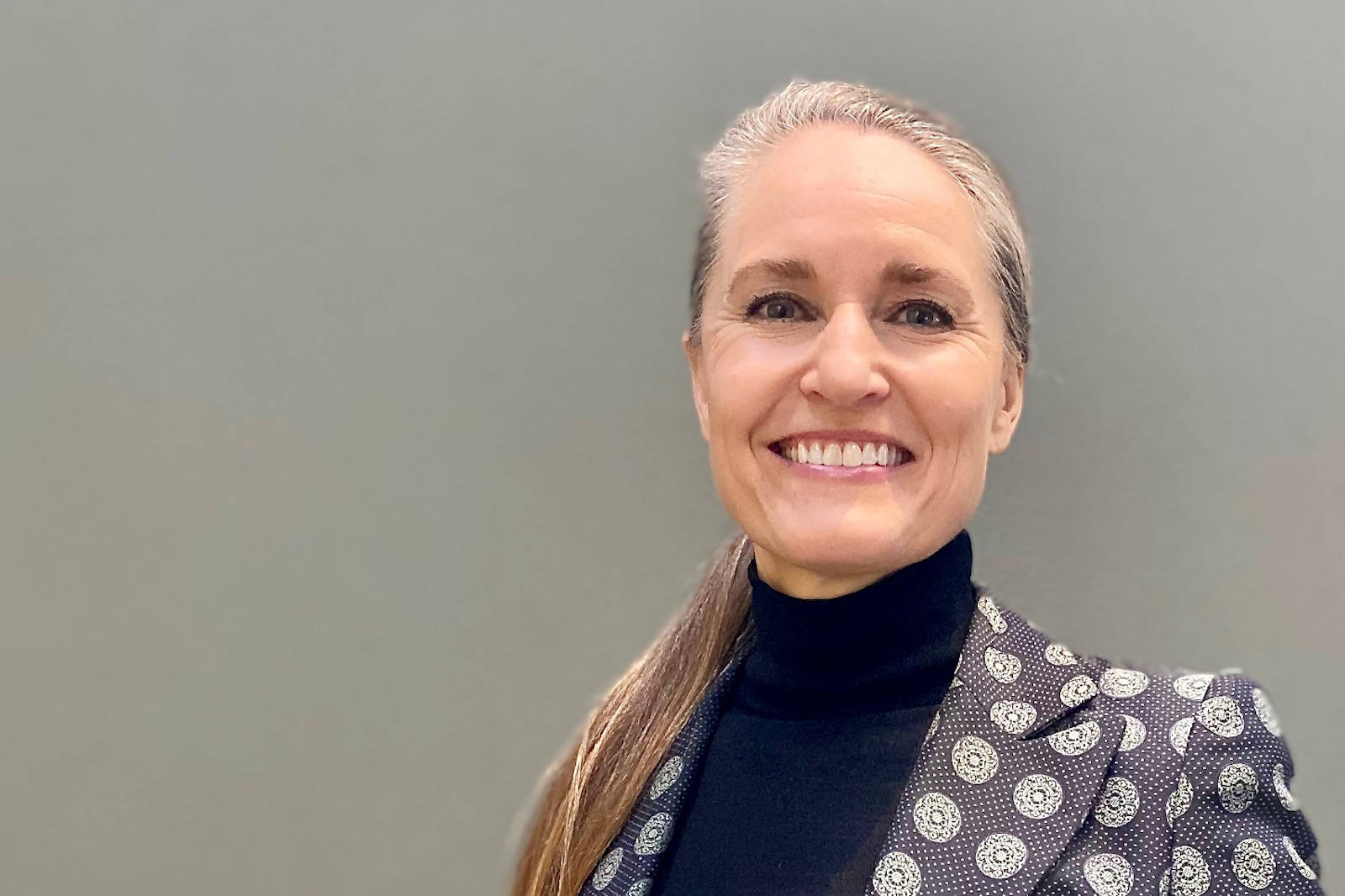 A person in an elegant polka dot suit stands against a grey background. Portrait of Helle Valentin.