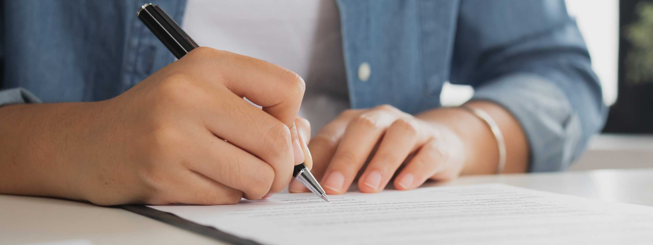 Eine Hand, die mit einem schwarzen Stift auf Papier schreibt, auf einem Tisch mit einem blauen Hemd im Hintergrund.