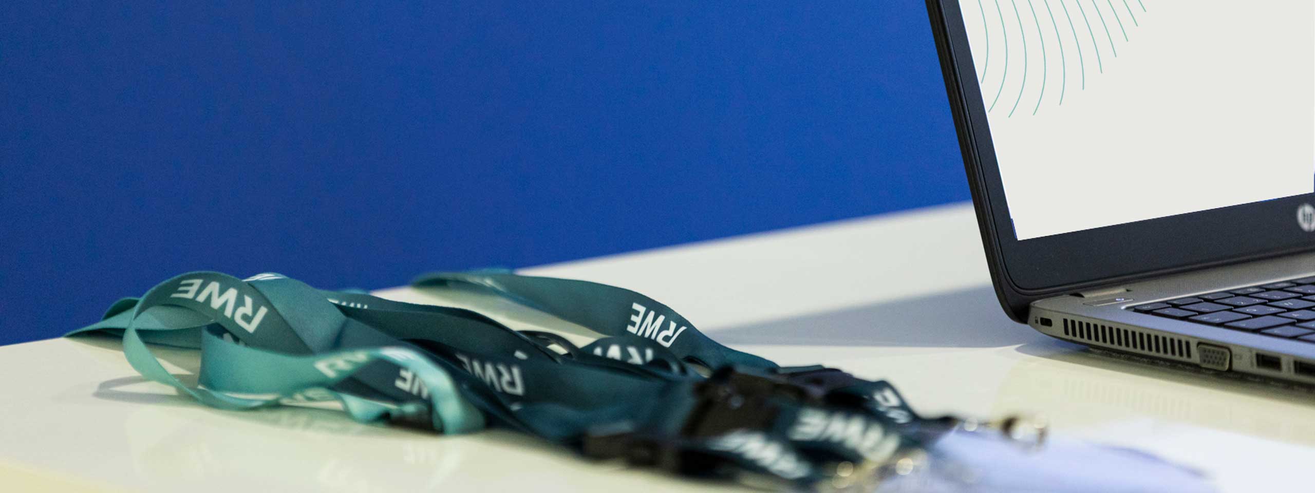 Eine Sammlung von türkisen Lanyards mit 'PRWE' darauf, neben einem Laptop auf einem weißen Tisch vor einem blauen Hintergrund.