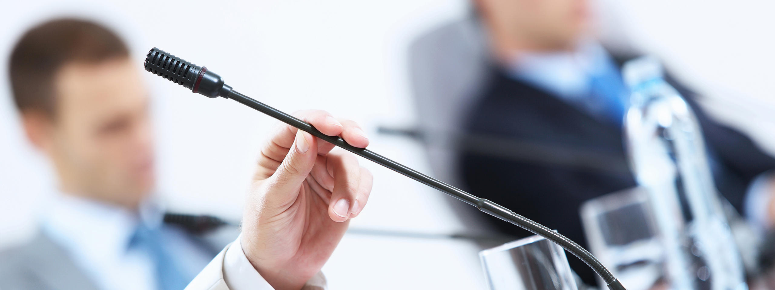 A hand holds a microphone while another person sits in the background. A glass of water is on the table.