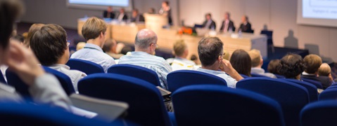 Audience members engaged in a conference session, facing a panel of speakers in a modern auditorium.