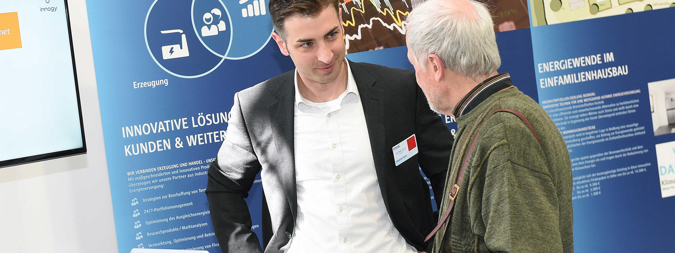 Two individuals discussing in front of a blue information stand about innovative energy solutions. A screen is visible.