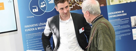 Zwei Personen im Gespräch vor einem blauen Informationsstand über innovative Energielösungen. Ein Bildschirm ist sichtbar.