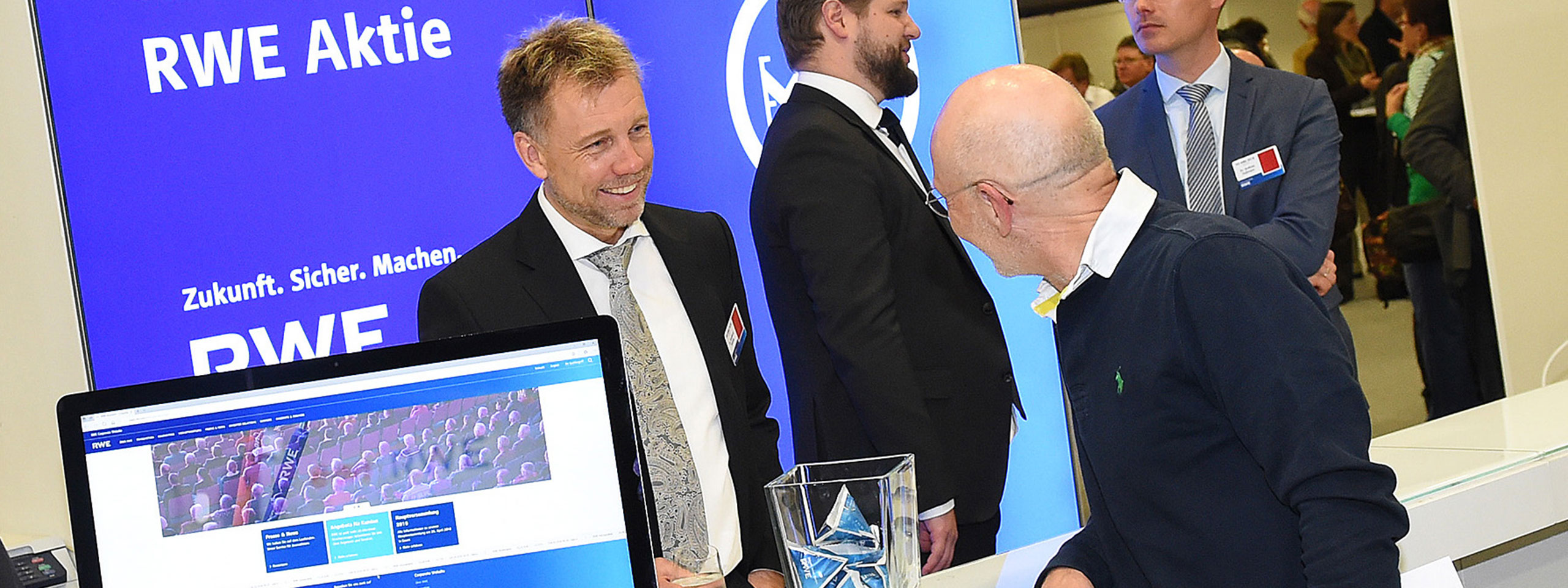 Messestand mit RWE Logo. Personen in Anzügen interagieren, ein Laptop zeigt die RWE-Website und eine Glasvase steht daneben.