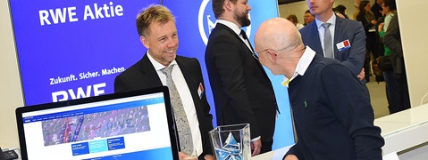 RWE booth featuring an RWE logo. People in suits interact, a laptop shows the RWE website, and a glass vase is nearby.