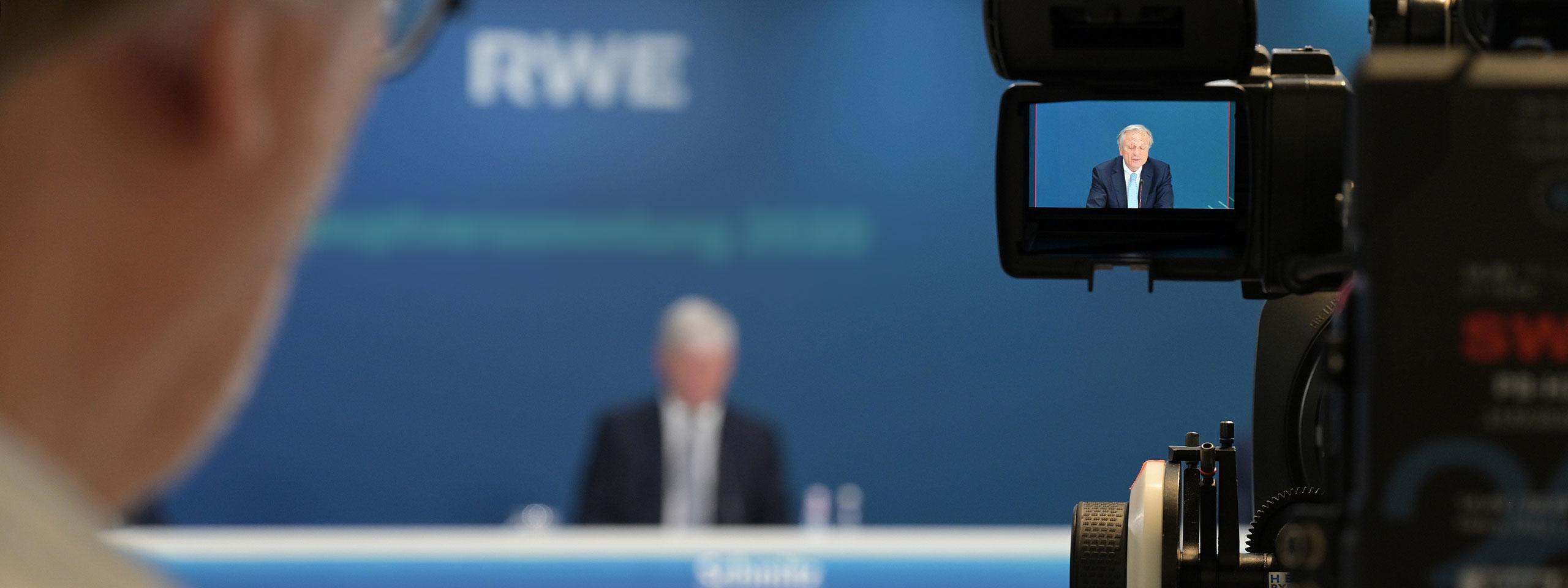 A camera captures a speaker at a conference, with a blue background displaying the RWE logo.