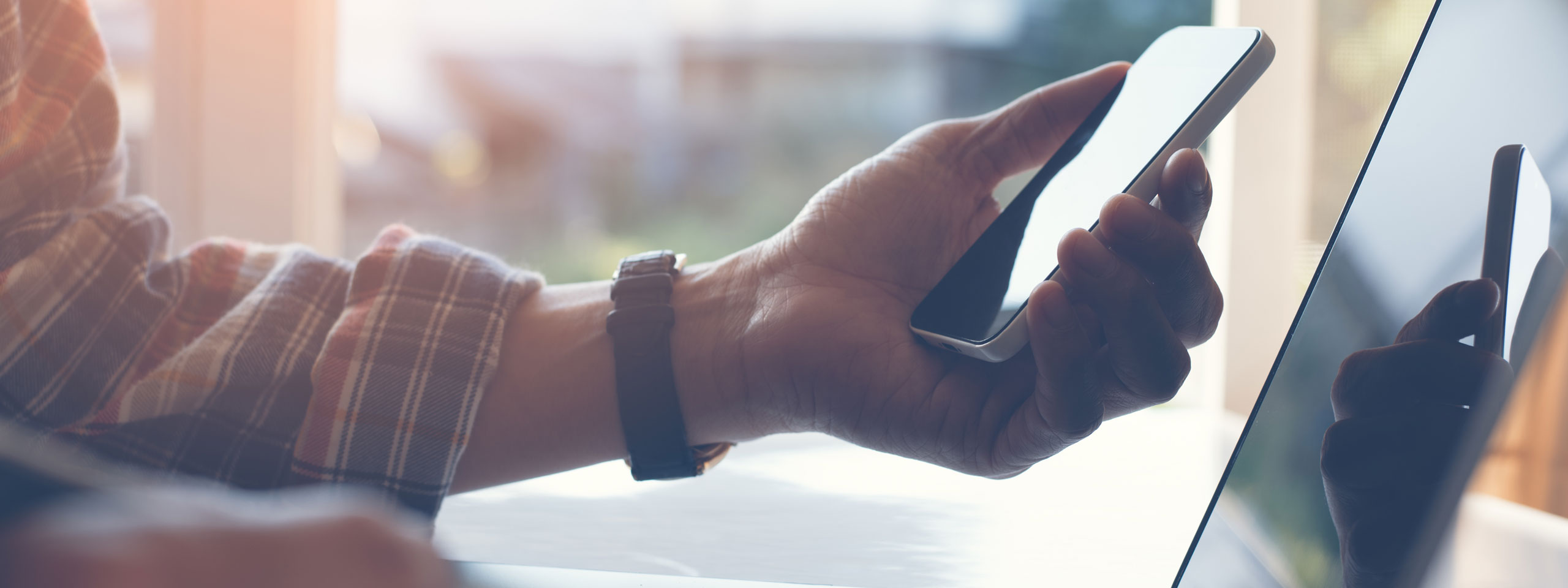 A hand holds a smartphone while looking at a laptop. A window in the background lets sunlight in.
