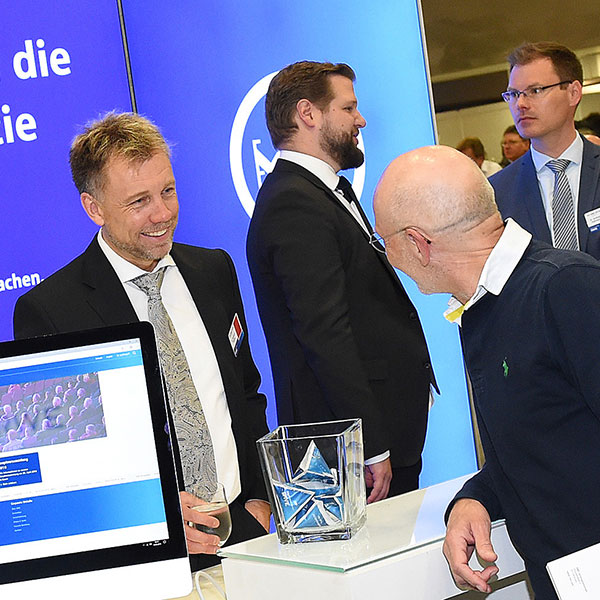 Eine Gruppe von Männern bei einem Messe-Stand, ein Computerbildschirm und ein Glasobjekt sind sichtbar.
