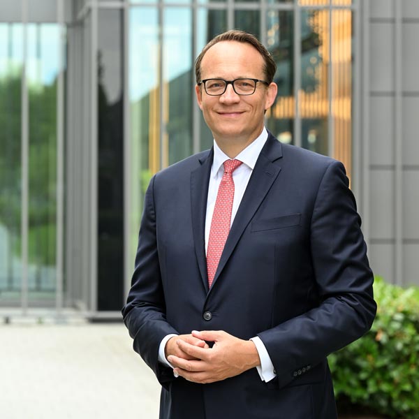 A man in a suit stands in front of a modern building. He has his hands in front and is wearing a red tie. Portrait of Markus Krebber.