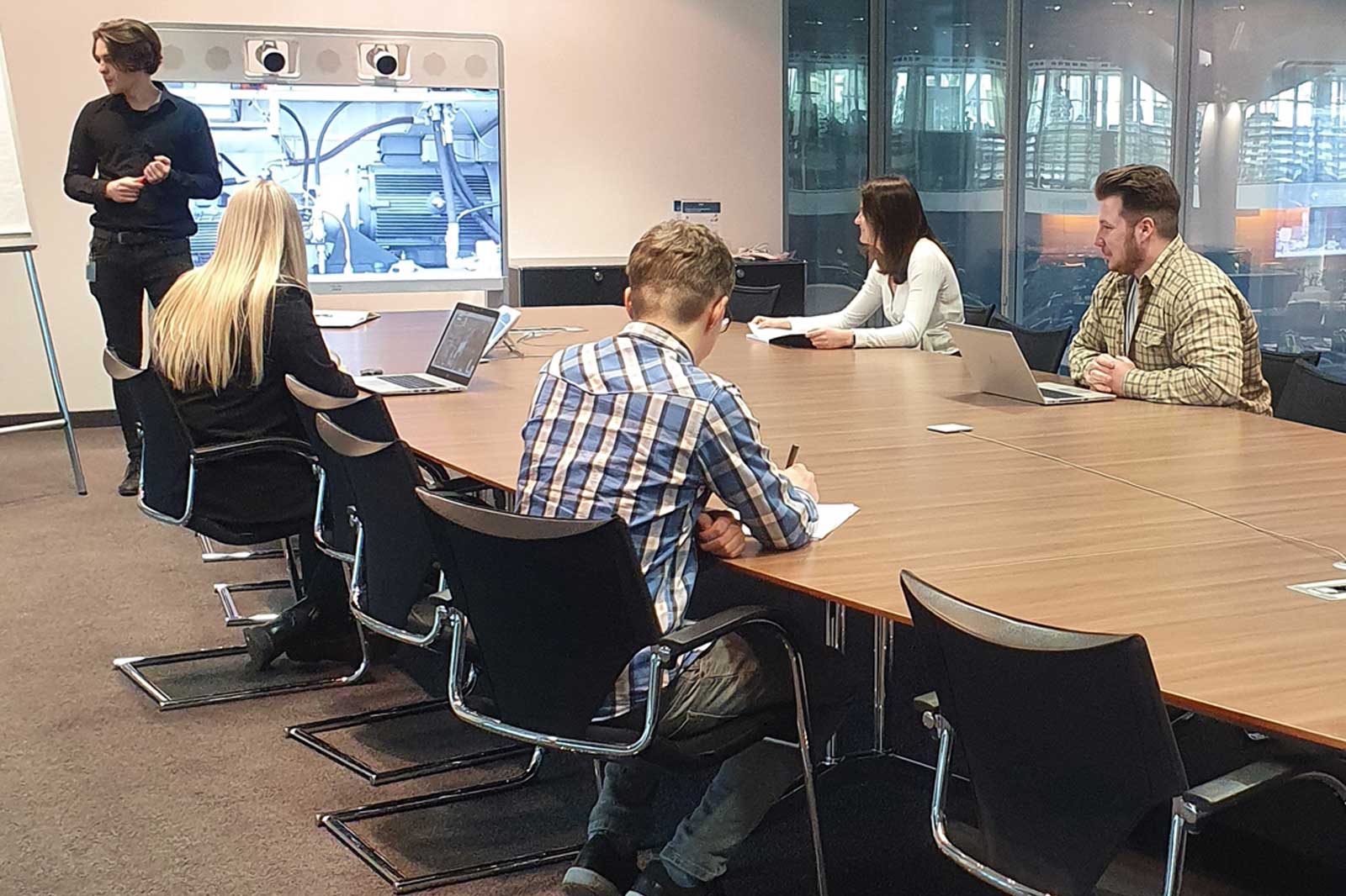 Vier Auszubildende zum Industriekaufmann (w/d/m) sitzen gemeinsam in einem Besprechungsraum und hören einem Kollegen bei einer Präsentation zu.
