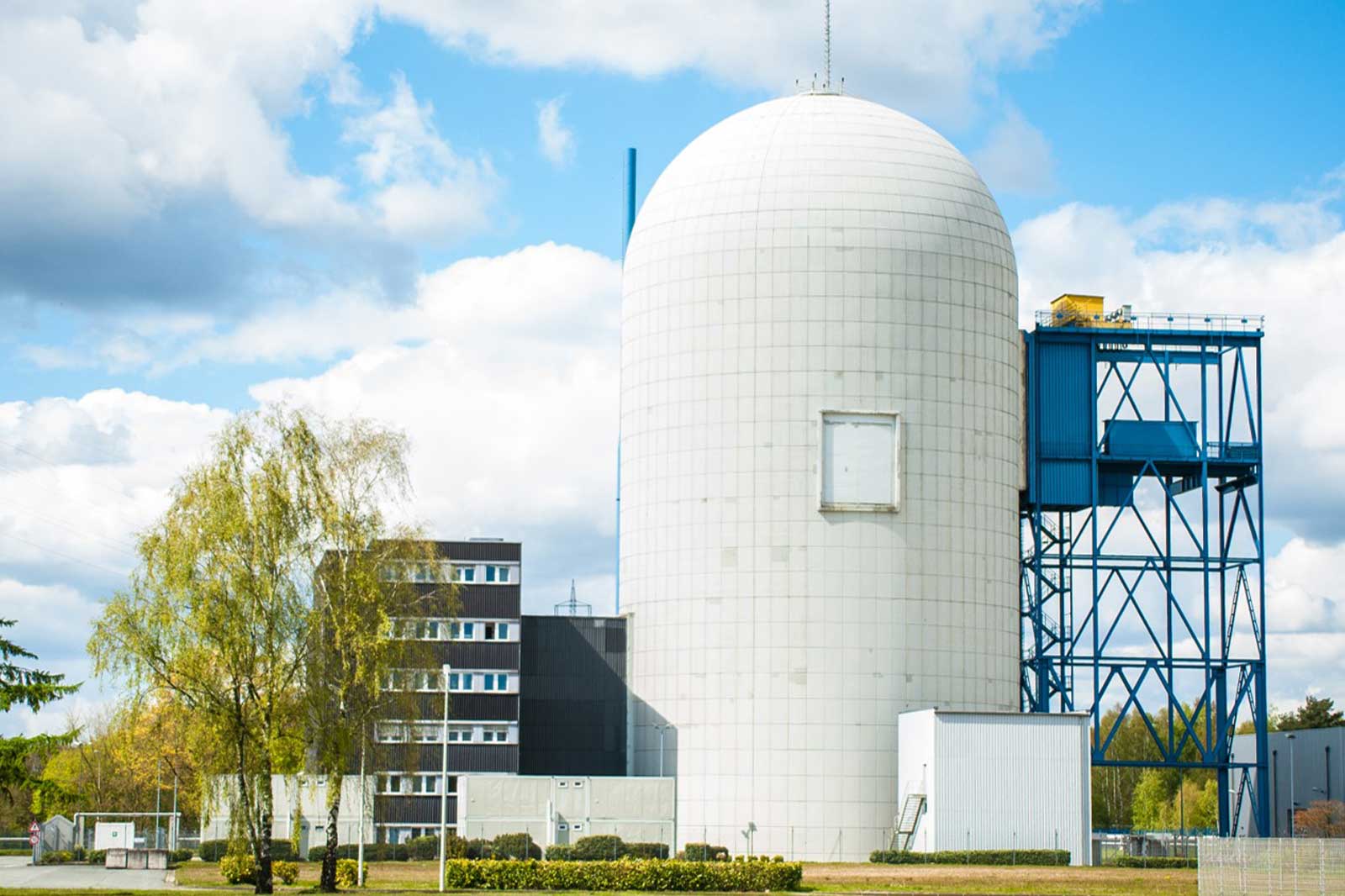 Eine große weiße zylindrische Struktur mit einem blauen Rahmen, begleitet von modernen Gebäuden und Grünflächen unter einem teilweise bewölkten Himmel.