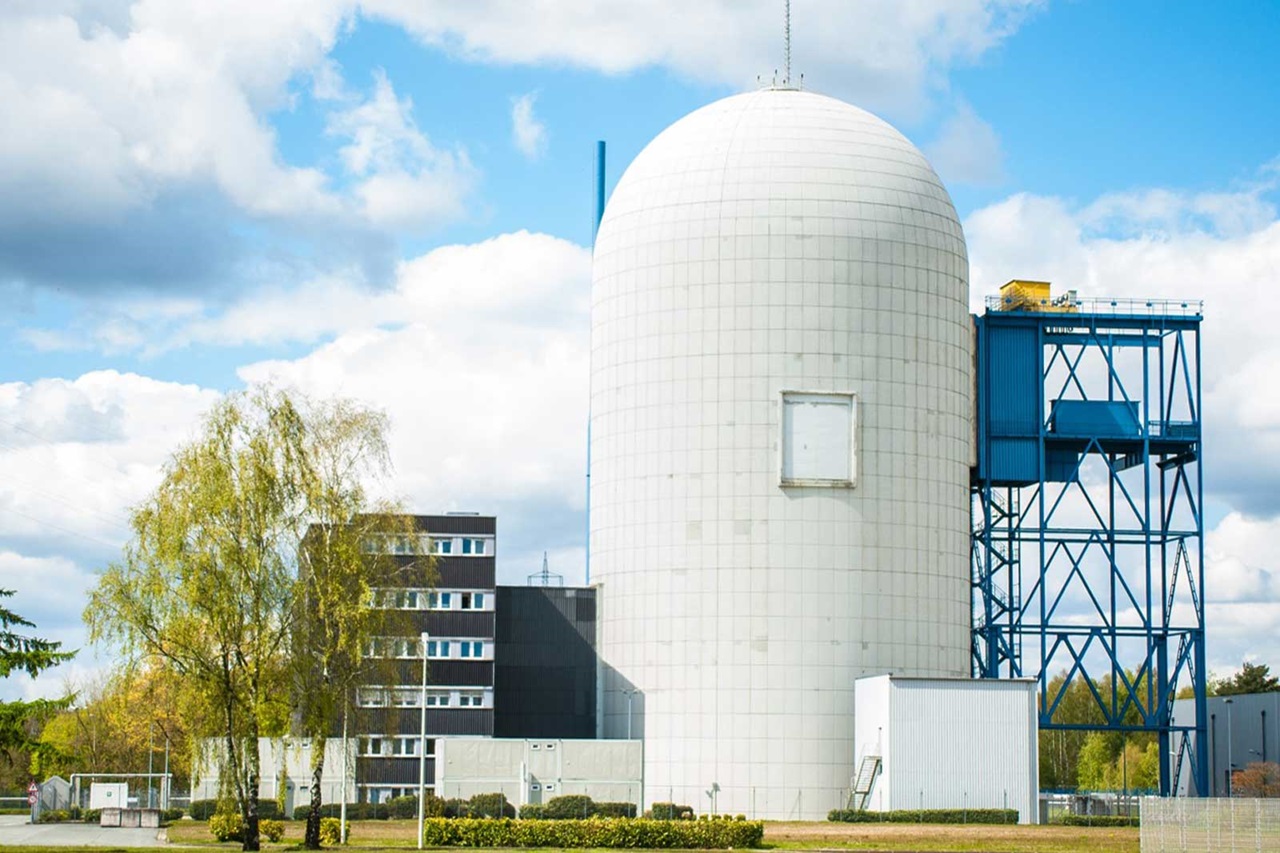 Eine große weiße zylindrische Struktur mit einem blauen Rahmen, begleitet von modernen Gebäuden und Grünflächen unter einem teilweise bewölkten Himmel.