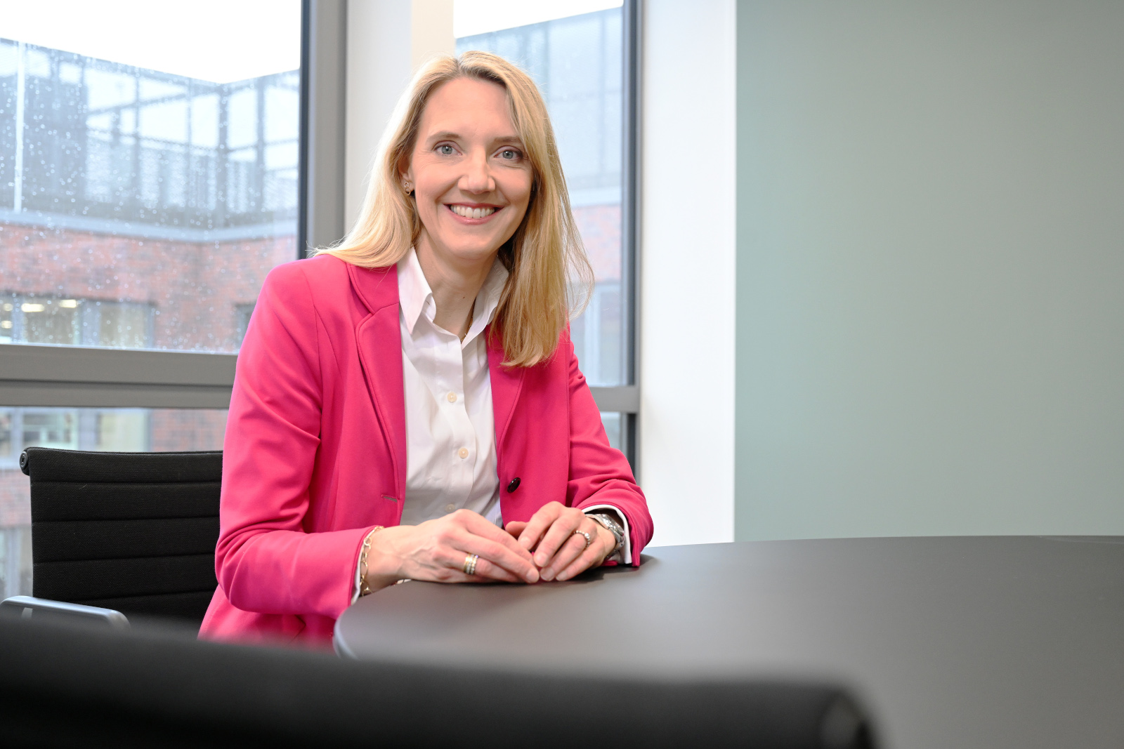 A woman in a pink blazer sits at a black table in a bright office with large windows and a green wall.
