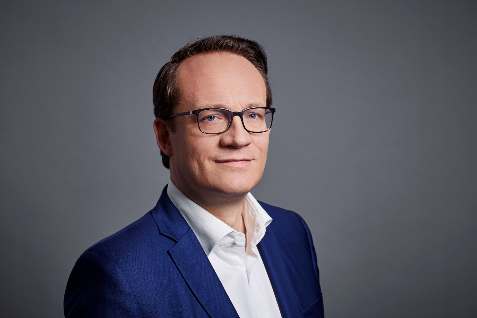 Ein Mann in einem blauen Anzug und weißem Hemd posiert vor einem neutralen Hintergrund.